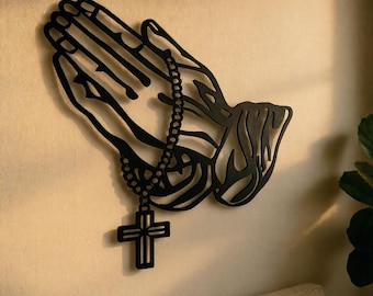 Może przedstawiać: Czarna metalowa dekoracja ścienna przedstawiająca modlące się dłonie trzymające różaniec z krzyżem. Dłonie są misternie wycięte, tworząc efekt cienia. Religijny element wystroju domu.