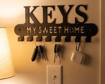 Może przedstawiać: Czarny metalowy wieszak na klucze z napisem "KEYS" nad "MY SWEET HOME". Z haczyków zwisa kilka kluczy i białe etui. Wieszak na klucze jest zamontowany na beżowej ścianie.