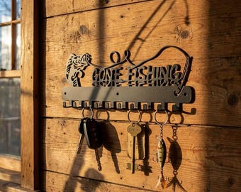 Może przedstawiać: Czarny metalowy wieszak na klucze z wyciętym napisem "GONE FISHING". Wieszak na klucze ma wzór wędki i rząd haczyków trzymających klucze i przynęty wędkarskie. Jest zamontowany na drewnianej ścianie.