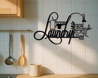Może przedstawiać: Czarna metalowa grafika ścienna z napisem "Laundry" w kursywie, z obrazami związanymi z praniem. Projekt obejmuje sznur do wieszania ubrań z odzieżą, pralkę i kosz. Grafika jest zamontowana na białej, kafelkowanej ścianie.