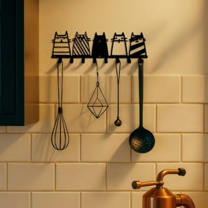 May include: Black metal wall-mounted kitchen utensil rack featuring five cat silhouettes. Below the cats are hooks holding a whisk, geometric pendant, small spoon, and slotted spoon. The rack is mounted on a tiled wall.
