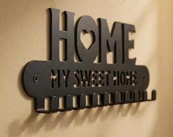 Może przedstawiać: Czarny metalowy wieszak na klucze z napisem "HOME" i wycięciem w kształcie serca w literze "O". Poniżej wyświetla się fraza "MY SWEET HOME". Wieszak na klucze ma wiele haczyków na klucze i jest zamontowany na beżowej ścianie.