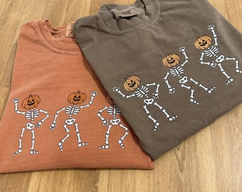 May include: Two crew neck sweatshirts, one orange and one brown, featuring a white skeleton design with orange pumpkin heads. The skeletons are in a dancing pose. The sweatshirts are folded on a wooden surface.