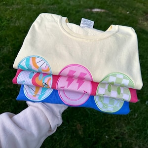 May include: A stack of four t-shirts with different smiley face designs. The top shirt is yellow, the second is pink, the third is blue, and the bottom is green. Each shirt has a different smiley face design, including a rainbow, lightning bolts, and a checkered pattern.