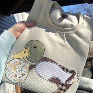 May include: A beige sweatshirt featuring an appliqued duck design. The duck has a green head, yellow beak, and a body composed of floral, striped, and plaid patterns. The sweatshirt is made of a soft material.