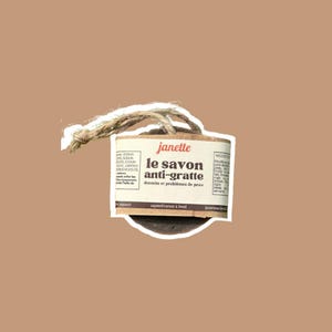 May include: A brown soap bar with a white label and a natural twine loop. The label reads "janette le savon anti-gratte dermite et problèmes de peau". The soap is a natural product.