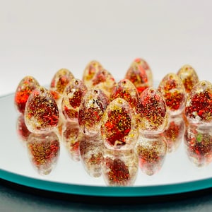 May include: A group of red and gold glitter filled glass beads in the shape of an egg. The beads are arranged in a circle on a reflective surface.
