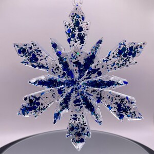 May include: A clear glass snowflake ornament with blue glitter embedded inside. The snowflake has six points and is suspended from a gold string.