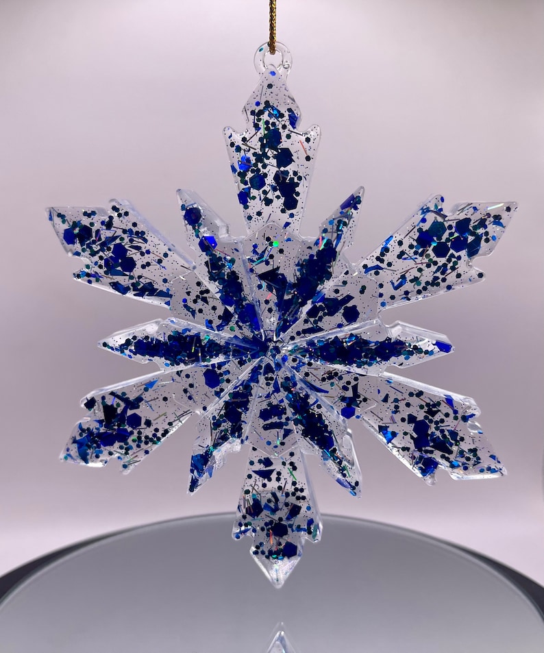 May include: A clear glass snowflake ornament with blue glitter embedded inside. The snowflake has six points and is suspended from a gold string.