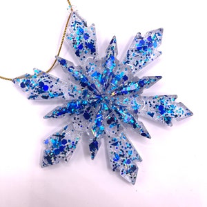 May include: A clear blue and silver snowflake ornament with glitter. The snowflake is made of resin and has a gold string for hanging.