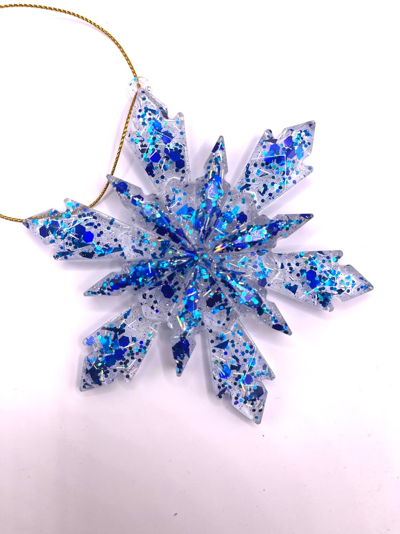 May include: A clear blue and silver snowflake ornament with glitter. The snowflake is made of resin and has a gold string for hanging.