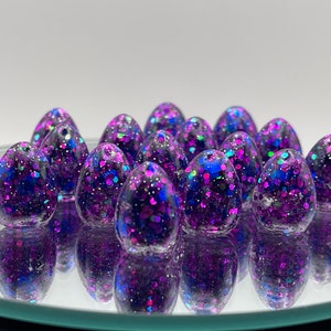 Puede incluir: Un grupo de 15 huevos de resina con purpurina de color morado y azul. Los huevos son pequeños y tienen un acabado brillante.