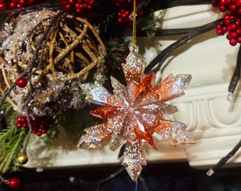 May include: A glittery, snowflake-shaped Christmas ornament with orange and silver accents. The ornament is suspended by a gold string and is surrounded by festive decorations, including red berries and greenery.
