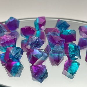 May include: A collection of 15 iridescent purple and blue gemstone-like resin dice with glitter. The dice are faceted and have a holographic finish.