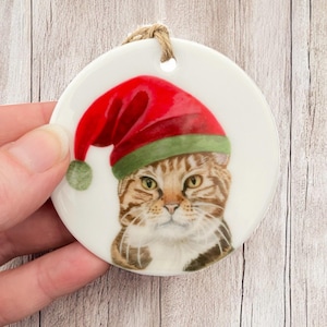 May include: A round, white Christmas ornament featuring a tabby cat wearing a red Santa hat with a green band and a small green pom-pom. The ornament has a jute string for hanging.