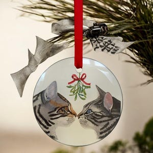 May include: A round Christmas ornament featuring two watercolor-style tabby cats nose to nose under a sprig of mistletoe. The ornament hangs from a red ribbon and is decorated with a silver bow and greenery.