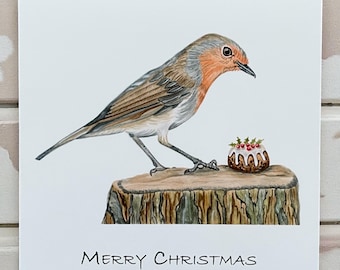 May include: A watercolor illustration of a robin perched on a tree stump next to a Christmas pudding. The bird has a red breast and brown wings. The words "MERRY CHRISTMAS" are written in black script at the bottom of the card.