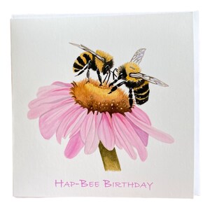 May include: A greeting card with a watercolor illustration of two bees on a pink flower. The card says "Hap-Bee Birthday" in pink lettering.