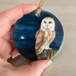 May include: Circular ceramic ornament depicting a barn owl on a wooden post against a dark blue night sky with a moon. The owl has white and brown feathers. A jute twine loop is attached for hanging.