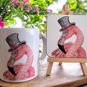 Pu&ograve; includere: Una tazza in ceramica bianca con un fenicottero rosa che indossa un cappello a cilindro nero. La tazza &egrave; accanto a un sottobicchiere di fenicottero rosa con un cappello a cilindro nero su un cavalletto di legno.