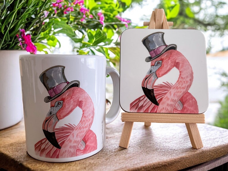 Pu&ograve; includere: Una tazza in ceramica bianca con un fenicottero rosa che indossa un cappello a cilindro nero. La tazza &egrave; accanto a un sottobicchiere di fenicottero rosa con un cappello a cilindro nero su un cavalletto di legno.