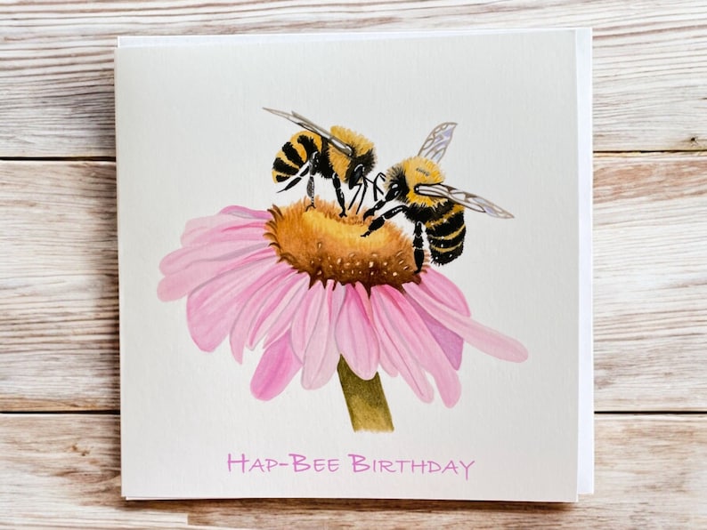 May include: A greeting card with a watercolor illustration of two bees on a pink flower. The card says "Hap-Bee Birthday" in pink lettering.
