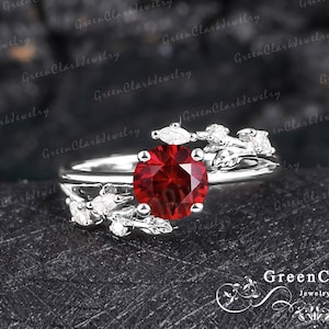 Könnte beinhalten: Ein silberner Ring mit einem roten Edelstein und einem filigranen Blattdesign. Der Ring liegt auf einem schwarzen Hintergrund.