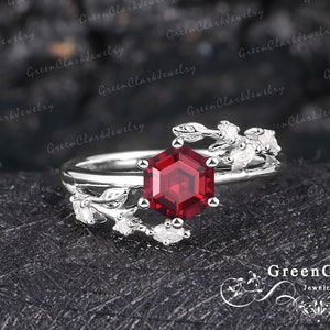May include: A silver ring with a hexagonal, deep red gemstone. The ring has a floral design with small white accents. The ring is set against a dark, textured background, highlighting the intricate details of the jewellery.