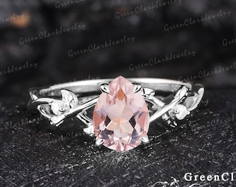 May include: A delicate silver ring with a pear-shaped pink gemstone set in a leafy design. The ring is on a black background.