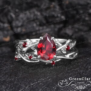May include: A silver ring with a pear-shaped, deep red gemstone as the centerpiece. The ring features a vine-like design with smaller red gemstones and is set against a dark, textured background. The ring is a piece of jewelry.