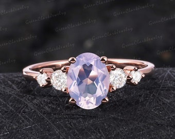 May include: Rose gold engagement ring with a large oval cut amethyst gemstone and smaller round cut diamonds on the band. The ring is on a dark gray textured surface.