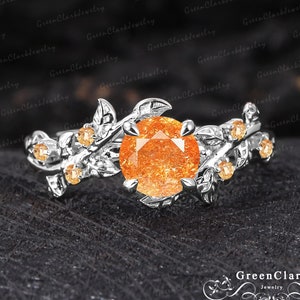 May include: A silver ring with a large orange gemstone surrounded by delicate leaves and smaller gemstones. The ring is set on a dark background.