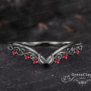 May include: A V-shaped ring featuring a dark metal band with intricate scrollwork and six small, round, vibrant red gemstones set along the curved design. The ring has a pointed centre and a polished finish, showcasing a detailed filigree pattern.