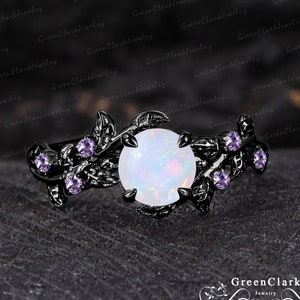 May include: A black metal ring featuring a large, round, white opal gemstone as the centerpiece. The ring is adorned with delicate leaf and vine detailing, accented by small, purple gemstones. The ring is set against a dark, textured background.