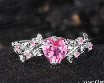 May include: A silver ring with a pink heart-shaped gemstone surrounded by delicate floral details. The ring features leaves and small pink gemstones.