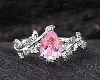 May include: A pear-shaped pink gemstone ring with a delicate silver band and green leaves. The ring is set on a black background.