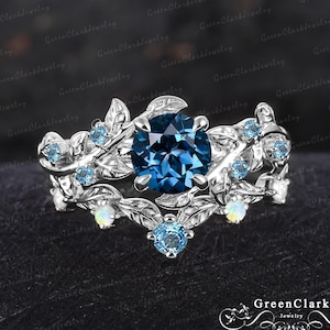 Op de afbeelding: Een zilveren ring met een blauwe edelsteen omringd door kleinere blauwe edelstenen en witte opaal accenten. De ring heeft een delicaat bladontwerp.