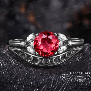 May include: A black metal ring set featuring a central round-cut red gemstone flanked by marquise-cut white stones. The band displays a decorative cutout pattern along the shank, and the text GreenClark Jewelry appears in the bottom right corner.