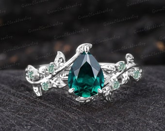 May include: A pear-shaped emerald engagement ring set in silver with a delicate vine design and small emerald accents. The ring is on a dark gray background.