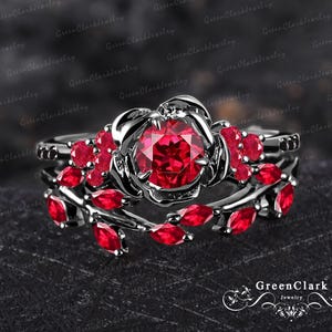 Könnte beinhalten: Ein schwarzer Rosen-Verlobungsring mit einem großen roten Rubin als Mittelstück und kleineren roten Rubin-Akzent-Steinen. Der Ring ist in einer schwarzen Metallfassung gefasst.