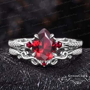 May include: A silver-toned ring set featuring a large pear-cut red centre stone, two smaller round red side stones, and a tiny red stone at the base. The band displays intricate scrollwork patterns and is accented with small clear stones.