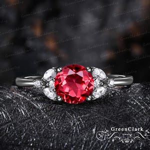 May include: A ring featuring a large, round, faceted red centre stone flanked by three smaller marquise-cut white stones on each side. The band is dark-toned metal, and the setting holds the stones in a cluster arrangement with a polished finish.