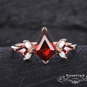 May include: Rose gold ring featuring a kite-shaped, deep red gemstone as the centerpiece, flanked by smaller, marquise-cut white gemstones. The ring has a delicate, floral-inspired design. The background is a textured, dark surface.