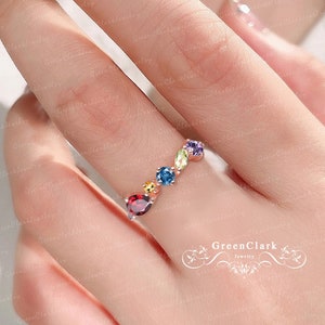 Könnte beinhalten: Ein roségoldener Ring mit einer Reihe farbiger Edelsteine. Der Ring zeigt einen tropfenförmigen roten Stein, gefolgt von gelben, blauen, grünen und violetten Steinen. Der Ring ist ein zartes Schmuckstück.