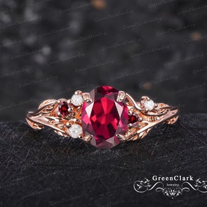 Può includere: Un anello di fidanzamento in oro rosa con una grande pietra preziosa di rubino rosso rotonda circondata da diamanti e rubini più piccoli. L'anello presenta un design delicato a forma di foglie.