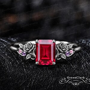 Op de afbeelding: Een zilverkleurige ring met een centrale smaragdgeslepen rode edelsteen. De band is aan beide zijden versierd met gebeeldhouwde metalen rozen en bladeren, geaccentueerd door kleine, ronde, lichtpaarse en roze steentjes in het bloemwerk.