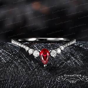 Peut inclure: Une bague noire avec un design incurvé, ornée d'une pierre rouge en forme de poire au centre. La bague est ornée de petits diamants blancs ronds et est posée sur un fond sombre. La bague est de GreenClarkJewelry.