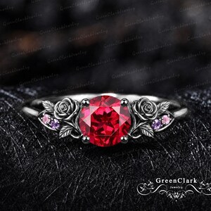 May include: A dark metal ring featuring a large, round, vibrant red centre stone. The band is decorated with sculpted rose motifs on both sides, accented by small, round, light pink and purple stones nestled within the floral metalwork.