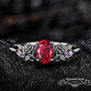 May include: A dark metal ring featuring a central oval-cut red gemstone. The band is decorated with sculpted rose motifs on both sides, accented by small round pink and purple stones nestled among the metallic leaves and petals.
