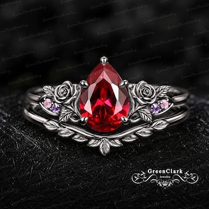 Peut inclure: Une bague en métal noir avec une pierre centrale rouge taille poire, entourée de motifs de roses sculptés et de petites pierres d'accent violettes et roses. L'anneau présente un design incurvé en forme de feuilles superposées.